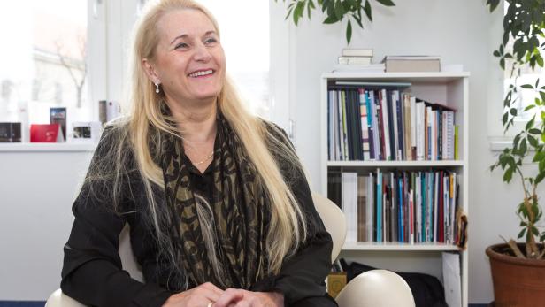 Eine lächelnde Frau mit blonden Haaren sitzt in einem hellen Raum vor einem Bücherregal.