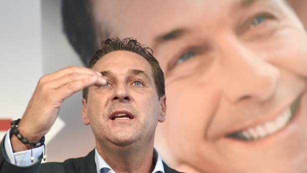 Ein Mann gestikuliert vor einem großen Porträt eines lächelnden Mannes.