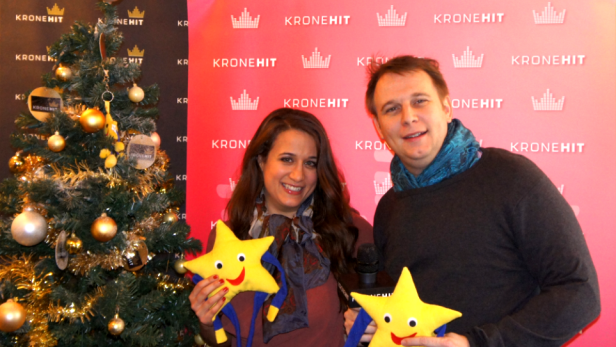 Eine Frau und ein Mann posieren mit Sternenpuppen vor einem Weihnachtsbaum und einem „Kronehit“-Hintergrund.