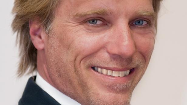 Ein lächelnder Mann mit blonden Haaren und blauem Hemd in einem dunklen Anzug.