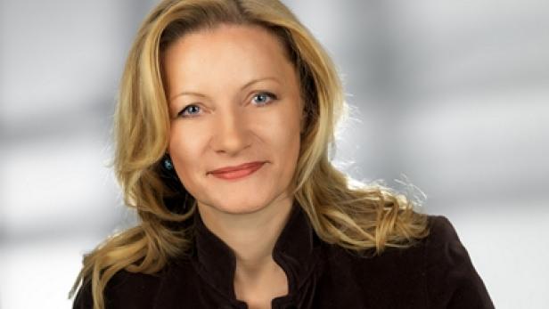 Porträt einer Frau mit blonden Haaren und brauner Jacke vor einem grauen Hintergrund.