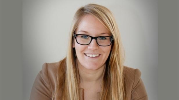 Eine lächelnde Frau mit blonden Haaren und Brille vor einem neutralen Hintergrund.