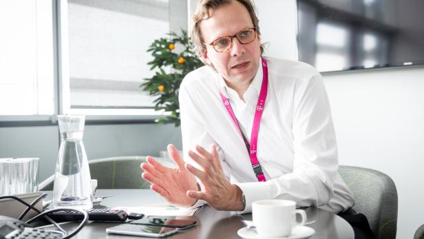 Ein Mann mit Brille und Telekom-Lanyard gestikuliert an einem Tisch mit Kaffeetasse.