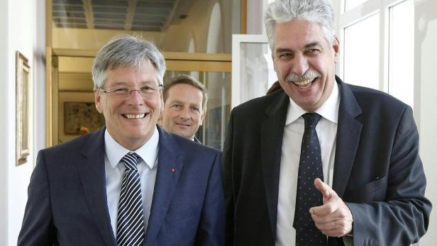 Zwei lächelnde Männer in Anzügen stehen in einem hellen Gebäude.