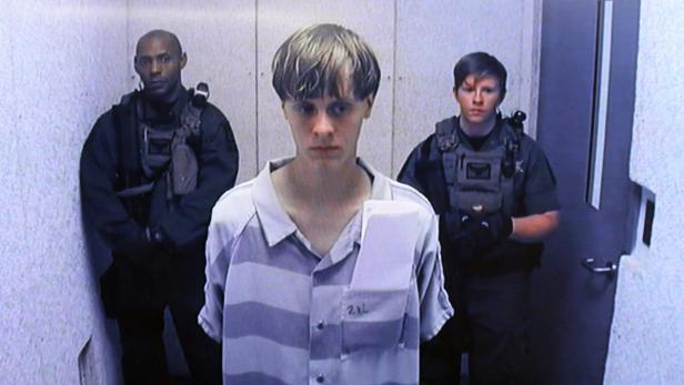 Dylann Roof in gestreifter Gefängniskleidung wird von zwei bewaffneten Beamten flankiert.