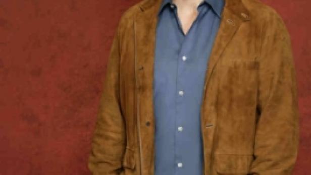 Nathan Fillion posiert vor einem roten Hintergrund in Jeans und einer Wildlederjacke.