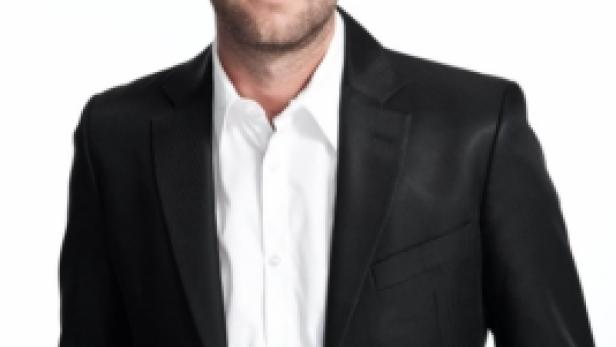 Ein Mann in einem schwarzen Anzug und einem weißen Hemd posiert vor einem weißen Hintergrund.