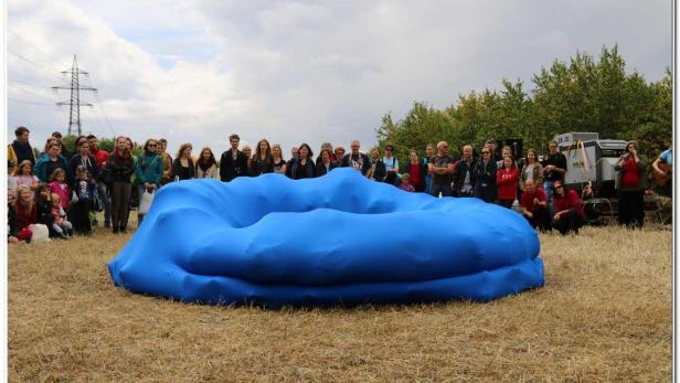 Eine Gruppe von Menschen steht um eine große, blaue, ringförmige Skulptur auf einem Feld.