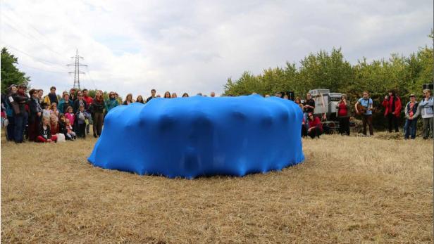 Eine Gruppe von Menschen steht um eine große, mit blauem Stoff bedeckte Struktur auf einem Feld.