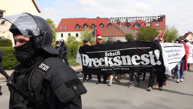 Ein Polizist steht im Vordergrund, während im Hintergrund eine Demonstration mit einem Banner stattfindet, auf dem „Scheiß Drecksnest!“ steht.
