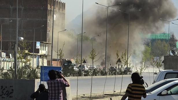 Eine Rauchsäule steigt in einer städtischen Umgebung auf, während Fotografen die Szene dokumentieren.