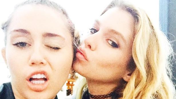 Miley Cyrus wird von einer Freundin auf die Wange geküsst.