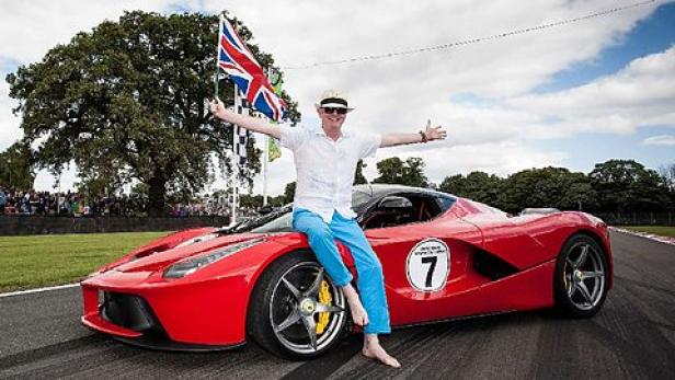 Ein Mann sitzt barfuß auf einem roten Ferrari mit der Nummer 7 und hält eine britische Flagge.