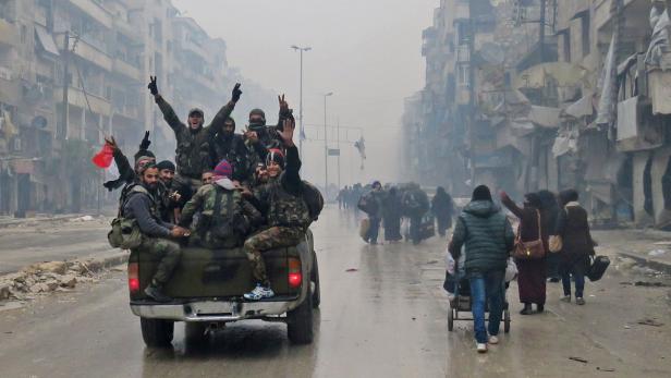 Bewaffnete Männer fahren auf einem Pickup durch eine zerstörte Stadt, während Zivilisten zu Fuß unterwegs sind.