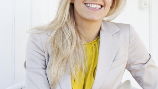 Eine lächelnde Frau mit blonden Haaren sitzt in einem Sessel.