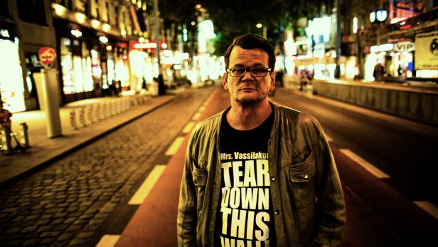 Ein Mann mit Brille und T-Shirt mit der Aufschrift „Tear Down This Wall“ steht nachts auf einer belebten Straße.