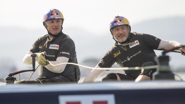 Zwei Segler mit Helmen von Red Bull Racing bedienen die Winden auf einem Segelboot.