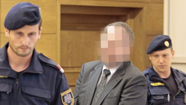 Ein Mann in einem Anzug wird von zwei Justizwachebeamten mit Baretten flankiert.