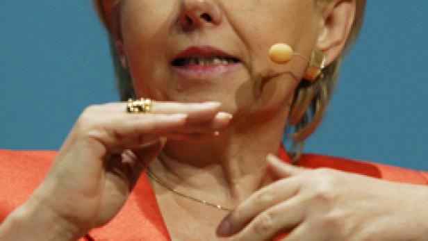 Eine Frau mit blonden Haaren und einem orangefarbenen Blazer spricht in ein Mikrofon.