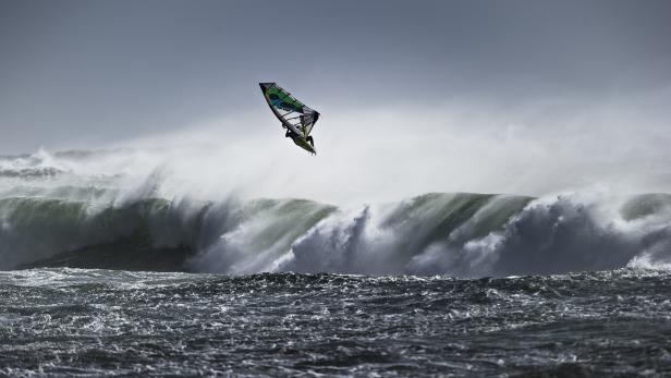 Ein Windsurfer springt vor einer großen Welle in die Luft.