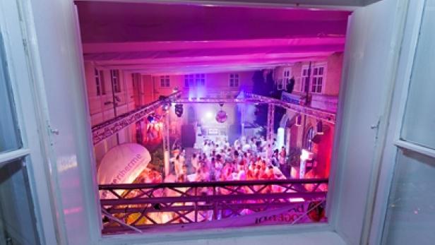 Blick aus einem Fenster auf eine Party mit vielen weiß gekleideten Menschen.
