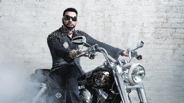 Ein Mann mit Sonnenbrille sitzt auf einem dekorierten Harley-Davidson-Motorrad vor einer Ziegelwand.