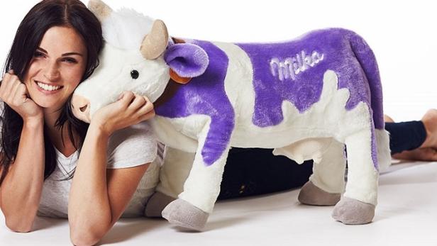 Eine Frau liegt neben einer großen, lila-weißen Milka-Kuh als Plüschtier.