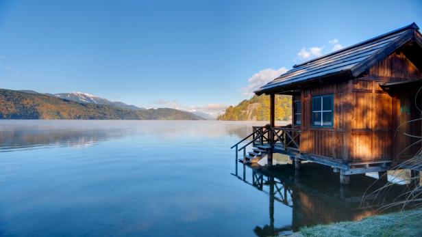 Ein Holzhaus steht auf Stelzen in einem ruhigen See mit Bergen im Hintergrund.