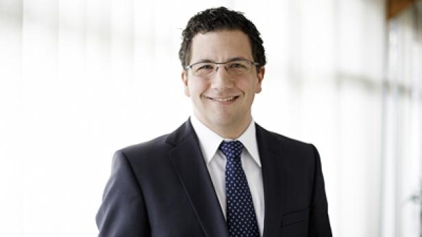 Ein lächelnder Mann mit Brille, Anzug und Krawatte vor einem hellen Hintergrund.