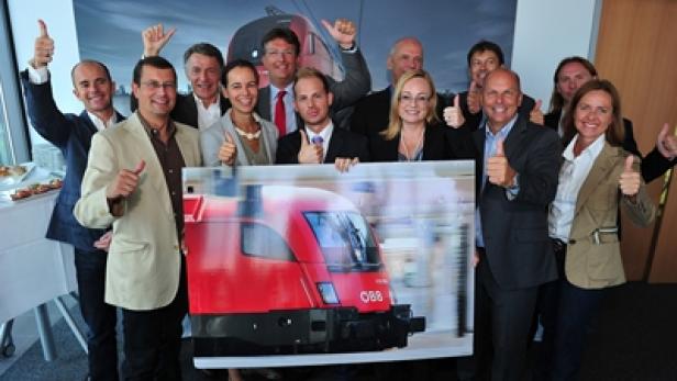 Eine Gruppe von Personen posiert mit Daumen hoch vor einem Bild eines ÖBB Railjet-Zuges.