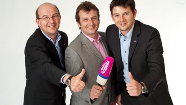 Drei Männer in Anzügen geben vor einem Mikrofon von Radio Arabella ein positives Zeichen.