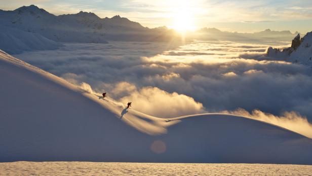 Zwei Skifahrer fahren einen schneebedeckten Hang hinunter, unter ihnen ein Wolkenmeer im goldenen Licht der Sonne.