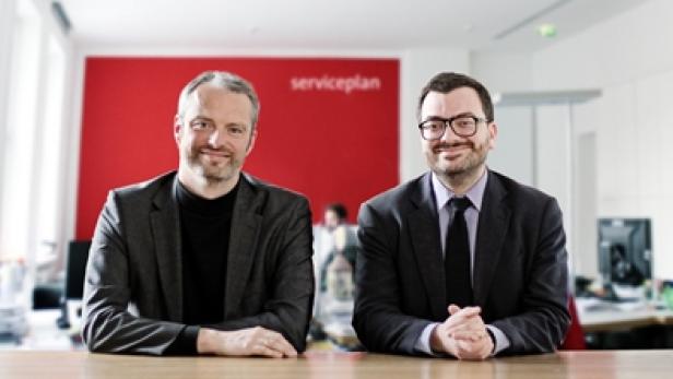 Zwei Männer sitzen an einem Tisch vor einer roten Wand mit der Aufschrift „Serviceplan“.