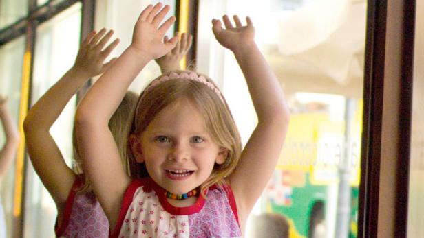 Ein Mädchen mit erhobenen Armen steht vor einem Fenster, hinter ihr weitere Kinder.