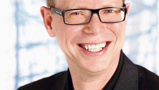 Ein lächelnder Mann mit Brille und dunklem Anzug vor einem hellen Hintergrund.