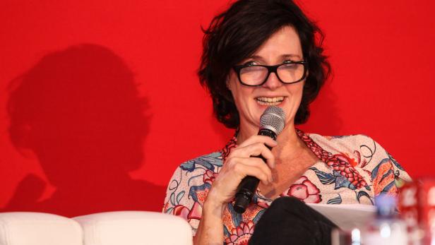 Eine Frau mit Brille spricht in ein Mikrofon vor einem roten Hintergrund.