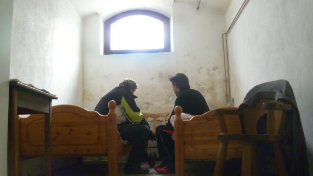 Zwei Personen sitzen in einem spärlich eingerichteten Raum auf Betten und unterhalten sich.