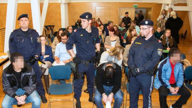 Drei Justizwachebeamte bewachen in einem Gerichtssaal drei mutmaßliche Täter mit Handschellen.