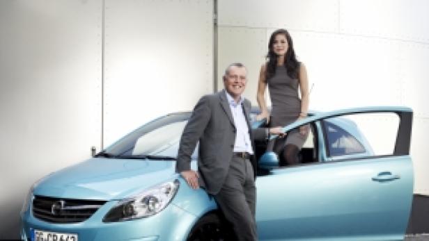 Ein Mann im Anzug und eine Frau posieren neben einem hellblauen Opel Corsa.