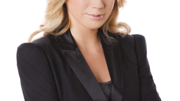 Eine blonde Frau in einem schwarzen Blazer mit verschränkten Armen vor einem weißen Hintergrund.