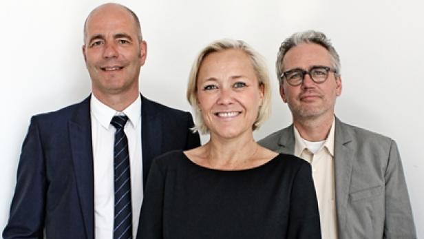 Drei Personen posieren vor einer weißen Wand für ein Gruppenfoto.