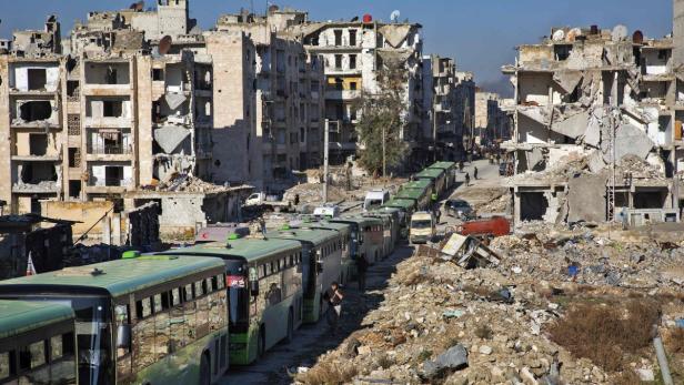 Eine Reihe grüner Busse steht vor zerstörten Gebäuden in einer Stadt.