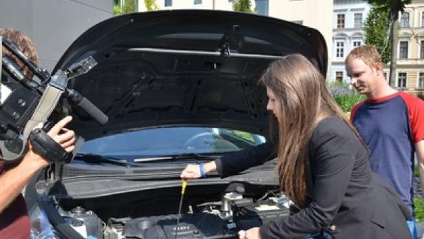 Eine Frau überprüft den Ölstand eines Autos, während ein Kameramann und ein Mann zusehen.