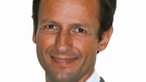 Ein lächelnder Mann in einem karierten Sakko vor einem weißen Hintergrund.