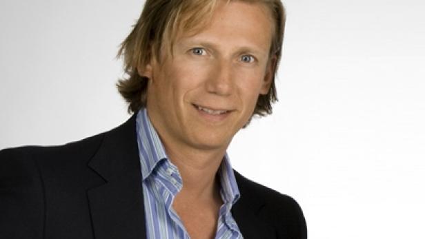 Ein Mann mit blonden Haaren trägt ein blau gestreiftes Hemd und ein dunkles Sakko.