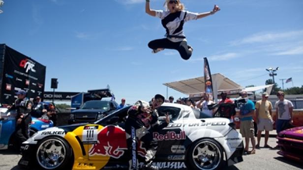 Ein Rennfahrer posiert mit einem Red Bull-Auto, während eine Frau in die Luft springt.