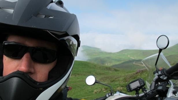 Ein Motorradfahrer mit Helm und Sonnenbrille vor einer grünen Hügellandschaft.