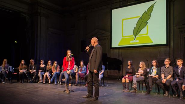 Eine Gruppe junger Leute sitzt auf einer Bühne, während ein Mann ins Mikrofon spricht. Im Hintergrund ist ein Bildschirm mit einem Laptop-Symbol zu sehen.