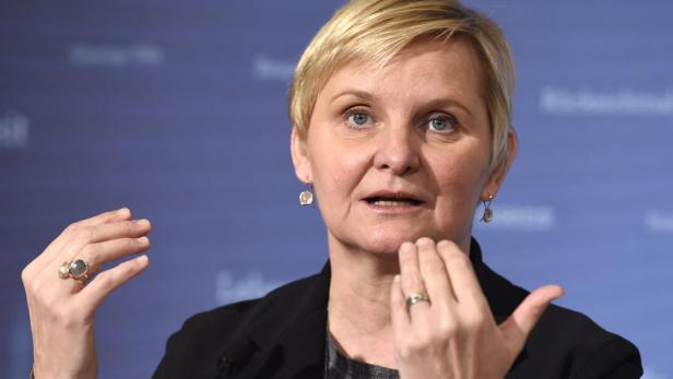 Eine Frau mit blonden, kurzen Haaren gestikuliert vor einem blauen Hintergrund.