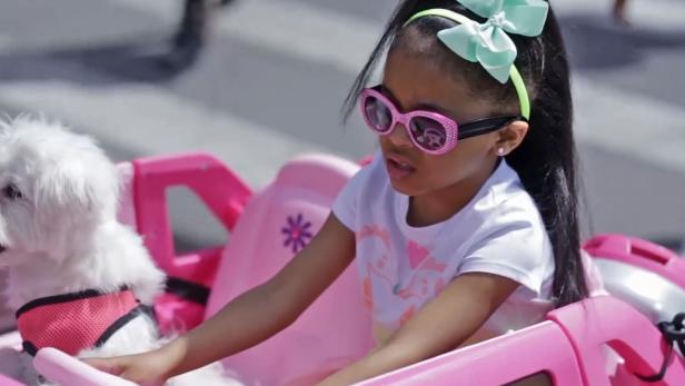 Ein Mädchen fährt mit einem kleinen, pinkfarbenen Auto, zusammen mit einem kleinen, weißen Hund.